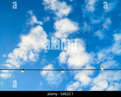 Blick aus dem niedrigen Winkel auf eine Reihe leuchtender Glühbirnen am blauen Himmel mit flauschigen weißen Wolken und Kopierbereich. Stockfoto