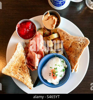 Komplettes nordamerikanisches Frühstück mit pochiertem Ei, Schinken, gebratenen Kartoffeln und Toast mit Erdnussbutter und Marmelade. Stockfoto