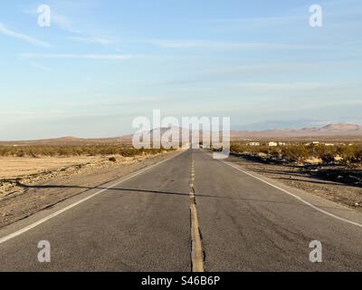 Wüstenstraße verschwindet in der Ferne, mit sichtbaren Bergen und dünnen Wolken am blauen Himmel über dem Himmel. Südkalifornien, am späten Nachmittag Stockfoto