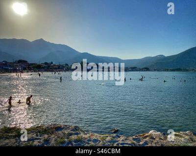 Menschen schwimmen bei Sonnenuntergang mit Blick auf die Hügel am Strand im Skala Potamias Resort auf der Insel Thassos, Griechenland. Stockfoto