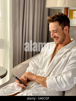 Der hübsche Mann im weißen Bademantel lächelt, während er sein Handy ansieht. Stockfoto