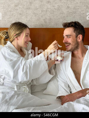 Junge kaukasische Paare in weißen Bademänteln gekleidet Ich bin ein Hotelzimmer Bett. Frau teilt ihr Essen mit ihrem Partner. Stockfoto