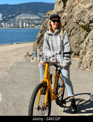 Junge blonde Hipster-Frau sitzt auf einem Fahrrad auf einem Strandweg, während sie nach rechts von der Kamera schaut. Stockfoto