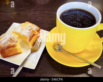 Kaffeepause. Ein langes Schwarz in einer gelben Tasse und Untertasse mit einer Scheibe glutenfreiem Orangenkuchen auf einem Teller auf einem Holztisch. Stockfoto
