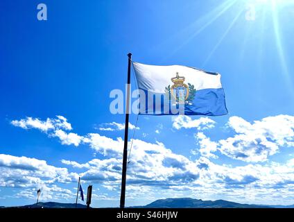 Die Flagge der Republik San Marion mit ihren hellblauen und weißen Bändern und ihrem nationalen Wappen in der Mitte weht vor dem blauen Himmel mit weißen Wolken und Sonnenstrahlen Stockfoto