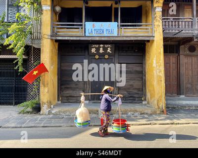 Vietnamese street vendor woman carrying goods in two baskets hanging at each end of a bamboo pole. Stock Photo
