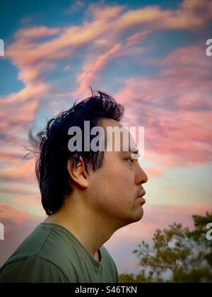 Porträt eines jungen asiatischen Mannes, der in der Betrachtung gegen rosa Wolken am Sonnenuntergangshimmel verloren ging. Stockfoto