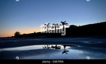 Weite Sicht, SUV-Silhouette und Palmen auf dem Hügel mit orangefarbenem Sonnenuntergangshimmel im Hintergrund und Reflexionen auf Pfützen auf dem Parkplatz im Vordergrund Stockfoto
