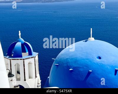 Die berüchtigte blaue Kuppel der Kirche Saint Spyridon und ihr Kuppelglockenturm, der von der Caldera in Oia, Santorin, Griechenland, flankiert wird. Stockfoto