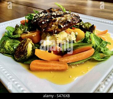 Marinierte gegrillte Hähnchenschenkel auf Ziegenkäse, Rucola, Spinat, geröstete Rosenkohl, gelbe Paprika, Karotten- und Tomatensalat, bedeckt mit einer asiatischen Sesamvinaigrette. Stockfoto