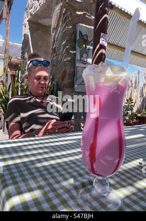 Eine Frau überprüft ihr Handy in einem Café im Freien mit einem bunten Getränk in Namibia Stockfoto