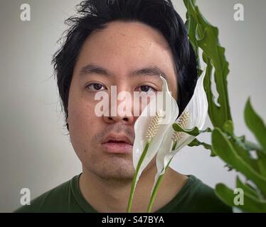 Nahaufnahme eines jungen asiatischen Mannes, eingerahmt von zwei Seerosenblüten und Philodendron-Blättern. Stockfoto