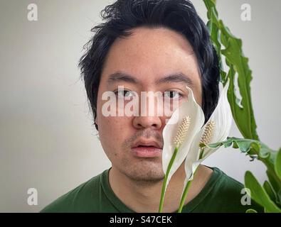 Nahaufnahme eines jungen, asiatischen Mannes, eingerahmt von zwei Seerosenblüten und Philodendron-Blättern mit Kopierraum. Stockfoto