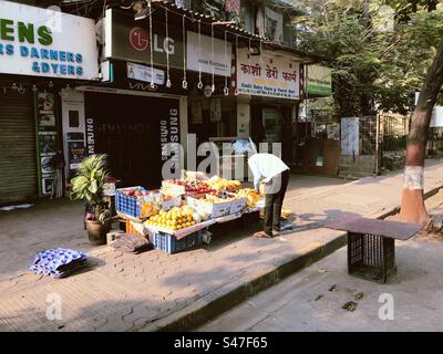 Ein Obstverkäufer auf einem Bürgersteig in Mumbai, Indien, mit Geschäften im Hintergrund Stockfoto