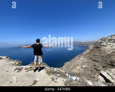 Junger Mann auf der vulkanischen Klippe der Insel Santorini mit Blick auf die Bucht von Anthinios und den Hafen von Athinios in der Ägäis. Stockfoto