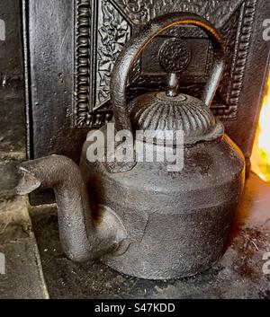 Ein viktorianischer gusseiserner Wasserkocher, der auf dem Kochfeld sitzt, ist eine alte Küchenzeile. Ein tosendes Feuer im Hintergrund. Stockfoto