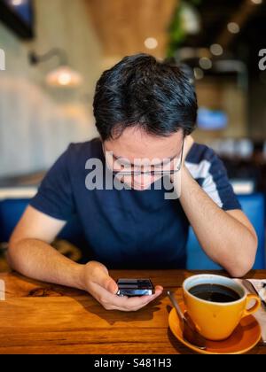 Junger asiatischer Mann mit Brille, der Handy benutzt, während er eine Tasse schwarzen Kaffee am Tisch in einem Café trinkt. Essen und Trinken. Wireless-Technologie. Fokus auf den Vordergrund. Stockfoto