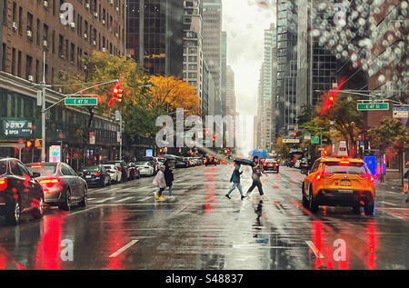 Regentag in New York auf der 47. Straße. Leute auf der Überfahrt mit Sonnenschirmen mit Autos, inklusive Taxi, die warten. Reflexionen auf nasser Straße von Brems- und Bremsleuchten Stockfoto