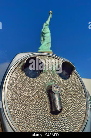 Nahaufnahme des Teleskops auf Liberty Island mit Freiheitsstatue im Hintergrund vor einem leuchtend blauen Himmel Stockfoto