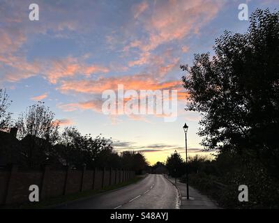 Sonnenuntergang über der Vorstadtlandschaft Großbritanniens. Blauer Himmel mit lachsrosa und orangen Wolken. Silhouettenbäume und Laternenpfosten. Stockfoto