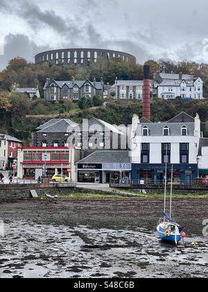 Ein blaues Segelboot im Oban Harbour, das bei niedrigem Wasser trocken steht. Mit einer Oban-Straße und dem Schornstein der Destillerie im Hintergrund. McCaig's Tower & Battery Hill Stockfoto