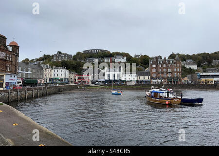 Ein Foto von Oban Harbour mit dem Schornstein der Destillerie und dem McCaig’s Tower im Hintergrund. Ein Segelboot und ein Touristenboot an der Front. Stockfoto