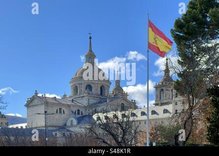 Eine spanische Flagge, die vor dem Königlichen Kloster El Escorial in Madrid, Spanien, fliegt. Ungewöhnliche Ansicht mit Kopierraum am blauen Himmel. Stockfoto