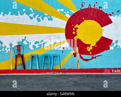 Wandgemälde mit großem rotem C, das oft auf der Flagge von Colorado zu sehen ist. Gelbe Sonnenstrahlen platzen aus. Roter Stuhl und 3 blaue Hocker stehen neben dem Gebäude und dem Parkplatz. Louisville, Colorado, USA. Stockfoto