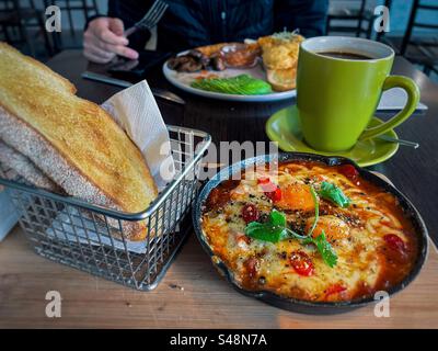 Shakshuka/pochierte Eier mit Bohnen in der Pfanne mit Sauerteig Toast im Korb und eine Tasse Kaffee gegen den mittleren Teil des Mannes, der die Gabel hält und das Handy in einem Café benutzt. Gesundes Frühstück. Stockfoto