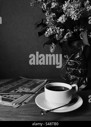 Kaffeetasse und Kreuzworträtsel-Zeitungen mit Fliederblüten in Schwarz-weiß Stockfoto