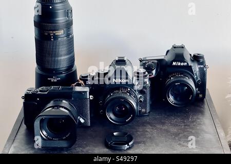 Studio-Aufnahme von drei Digitalkameras auf einem Holzregal. Ein mit Klebeband versehener Fujifilm X-E2s, ein Nikon DF und ein Nikon D780 mit verschiedenen Objektiven und Teleobjektiven. Stockfoto