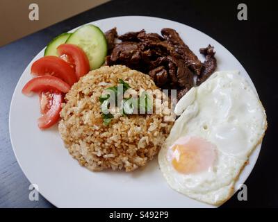 Ein weißer Porzellanteller mit hausgemachtem gebratenem Reis, sonniger Eierseite nach oben, geschnittenem Grillfleisch im asiatischen Stil, Gurken- und Tomatenscheiben auf einem schwarzen Holztisch. Stockfoto