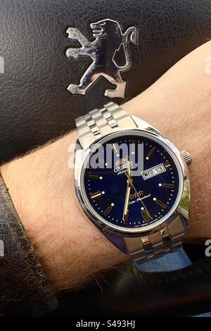 Nahaufnahme einer Orient Automatic Tristar Uhr mit tiefblauem Zifferblatt und goldenen Indexen und Händen am behaarten Handgelenk mit Lederjacke über einem schwarzen französischen Autolenkrad. Stockfoto