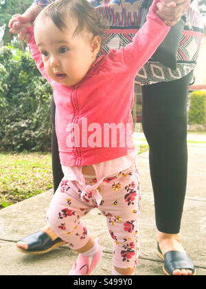 Die ersten Schritte Ihres Babys im Freien. Das Baby läuft mit Aufregung und wird von einem Erwachsenen unterstützt Stockfoto