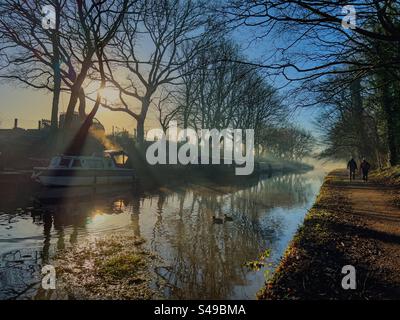 Reflexionen im Morgennebel des Winters auf dem Leeds- und Liverpool-Kanal in Adlington, Lancashire. Zwei Personen laufen auf einem Schleppweg mit Reflexen von Bäumen und schmalen Booten im Kanal. Stockfoto