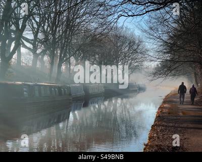 Zwei Personen laufen auf dem Schleppweg von Leeds und Liverpool Kanal im Morgennebel. Schmale Boote und Bäume spiegeln sich im Kanal. Adlington bei Chorley in Lancashire. Stockfoto
