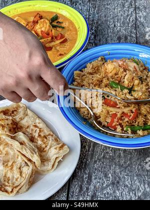 Löffel und Gabel mit der Hand über gebratenem thailändischem Basilikumreis in einer Schüssel mit rotem thailändischem Curry in einer Schüssel und Roti, gebratenem Fladenbrot auf dem Teller auf dem Tisch. Thailändisches Essen. Stockfoto