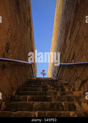 Niedriger Blickwinkel, Blick auf eine Steintreppe auf ein kleines Mädchen, eingerahmt am blauen Himmel Stockfoto