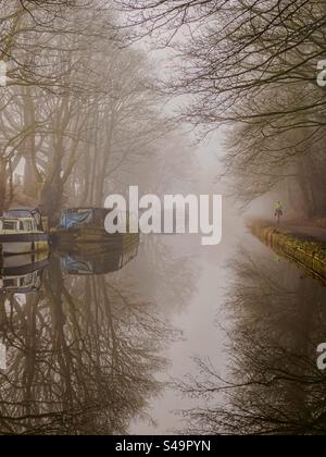 Läufer auf dem Schleppweg des Leeds- und Liverpool-Kanals an einem nebeligen Morgen mit Bäumen und schmalen Booten, die sich im Wasser spiegeln. Adlington in der Nähe von Chorley bin Lancashire. Stockfoto