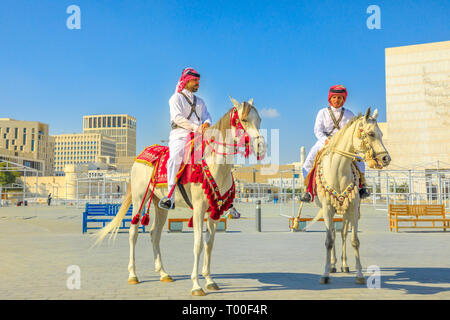 Doha, Katar - Februar 20, 2019: Zwei Erbe Polizeibeamte in traditionellen 1940 s Qatari einheitliche Reiten weiße Arabische Pferde bei Square von Souq Waqif Stockfoto