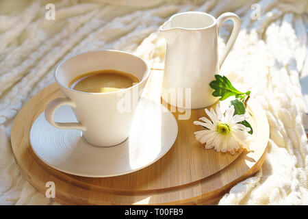 Weiße Tasse Kaffee mit Milch und Blume auf einem runden Holz- Fach auf gemütlichen gestrickt Plaid in sanften Morgensonne. Stockfoto