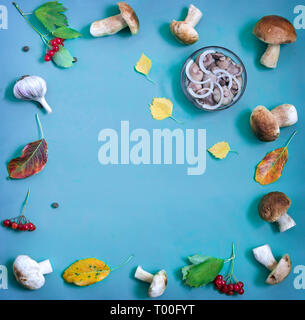 Auf einem blauen Hintergrund sind Pilze in einer Vase und in einem Glas marinierte, neben frische Pilze und Blätter im Herbst. Stockfoto