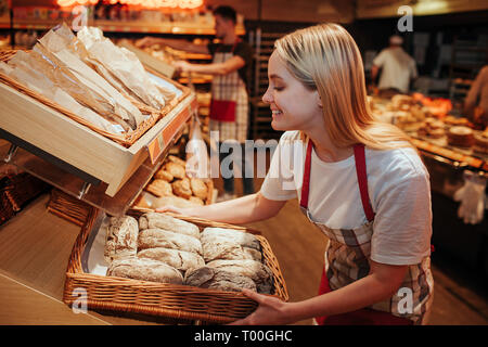 Junge Frau halten bakset mit frischem Brot im Supermarkt. Sie setzte es im Regal und Lächeln. Lecker und Delisious. Im Inneren arbeiten. Warmes Licht. Stockfoto
