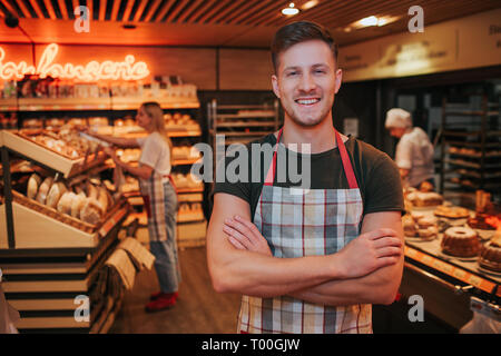 Junge starke gut aussehender Mann stand im Lebensmittelgeschäft oder Bäckerei. Er kreuzte die Hände und Lächeln. Positive Arbeiter. Junge Frau hinter in der Nähe der Bäckerei Regal Stockfoto