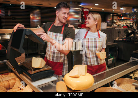 Junger Mann und Frau stand auf der Käse Regal im Supermarkt. Sie schauen sich an und lächeln. Frau halten Stück Käse in der Hand. Kerl stand auf der Stockfoto