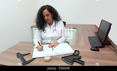 Weibliche latin Ärztin sitzen mit ernsten Einstellung in Ihrem Büro mit Stethoskop an ihrem Hals schreiben in ihrem Notebook auf weißem Hintergrund Stockfoto