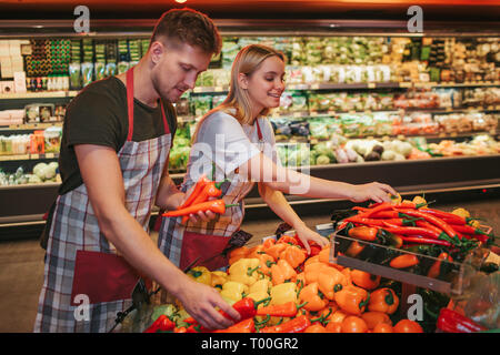 Junger Mann und Frau stand auf Gemüse Warenkorb im Lebensmittelgeschäft. Sie Paprika zusammen und Lächeln. Fleißige Leute. Stockfoto