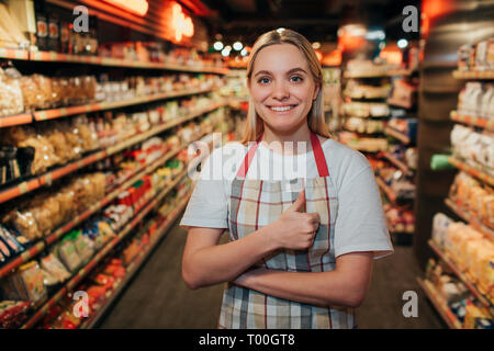 Happy positive junge Frau stand in line in Lebensmittelgeschäft. Sie schauen auf Kamera und Lächeln. Modell halten grosse Daumen nach oben. Stockfoto