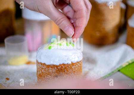 Baker schmückt den oberen Teil von Ostern Kuchen mit weißen Zucker durch farbige Kristallzucker gießen Dekoration auf frisch Puderzucker Puderzucker bedeckt, tra Stockfoto