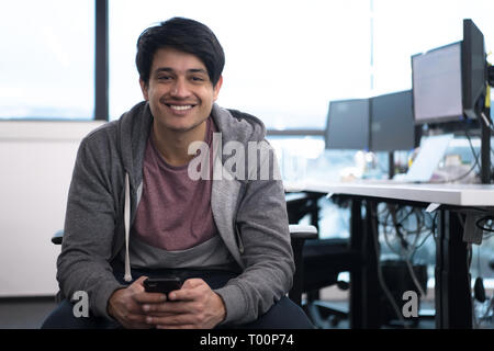 Junge indische männlichen Software Entwickler mit Handy Nachrichten schreiben, während Sie Pause an modernen, kreativen Start Büro Stockfoto